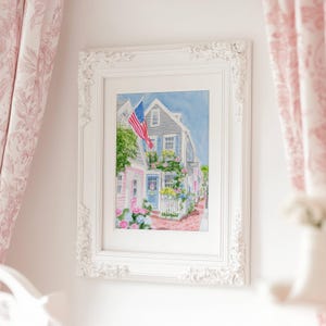 May include: A framed watercolour painting depicting a charming coastal town. The artwork shows a house with an American flag, surrounded by flowers and a white picket fence. The frame is white with decorative detailing.