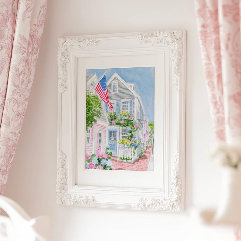 May include: A framed watercolour painting depicting a charming coastal town. The artwork shows a house with an American flag, surrounded by flowers and a white picket fence. The frame is white with decorative detailing.