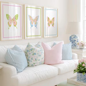 May include: Three framed butterfly art prints with gold frames and colorful watercolor designs hang above a white sofa. The sofa is adorned with striped and floral pillows, and a solid pink pillow. A lamp and flowers complete the room.
