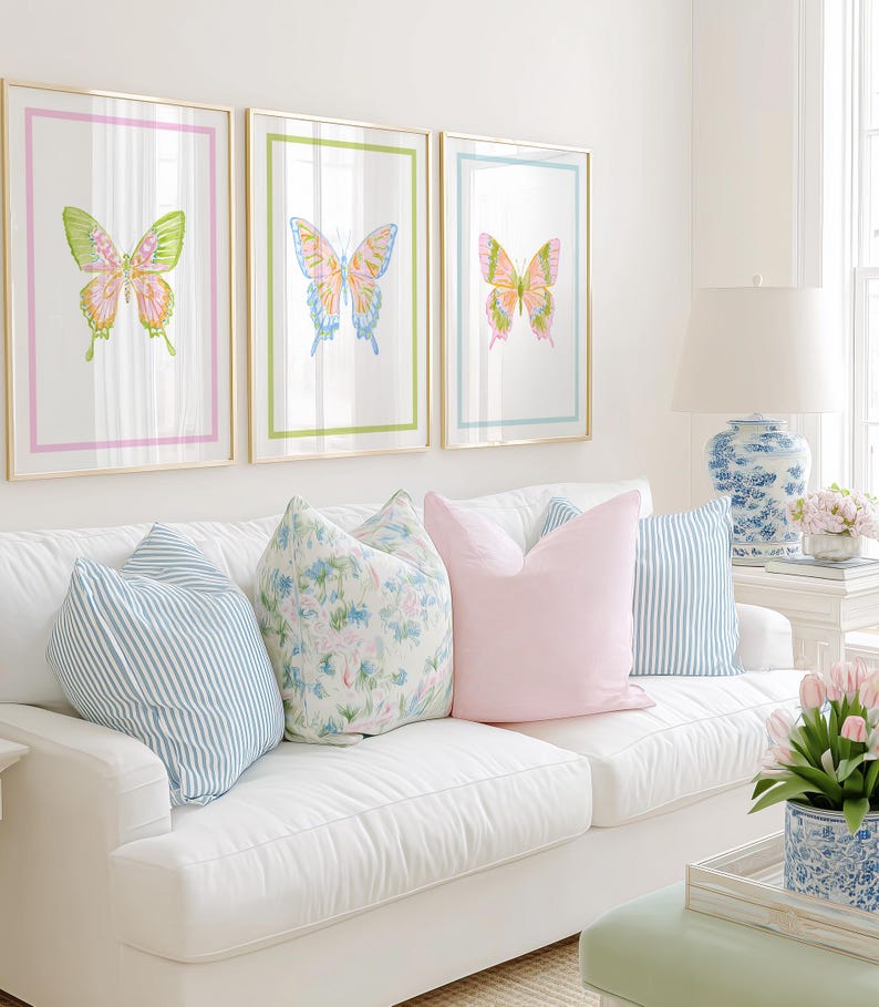 May include: Three framed butterfly art prints with gold frames and colorful watercolor designs hang above a white sofa. The sofa is adorned with striped and floral pillows, and a solid pink pillow. A lamp and flowers complete the room.