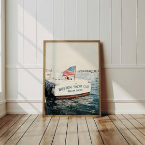 May include: Framed print of a white boat with the text "BOSTON YACHT CLUB MARBLEHEAD" and an American flag. The boat is docked in blue water. The print is in a wooden frame and leans against a white wall and wooden floor.