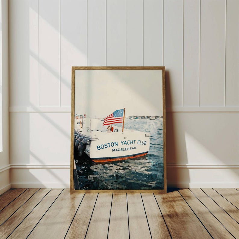 May include: Framed print of a white boat with the text "BOSTON YACHT CLUB MARBLEHEAD" and an American flag. The boat is docked in blue water. The print is in a wooden frame and leans against a white wall and wooden floor.