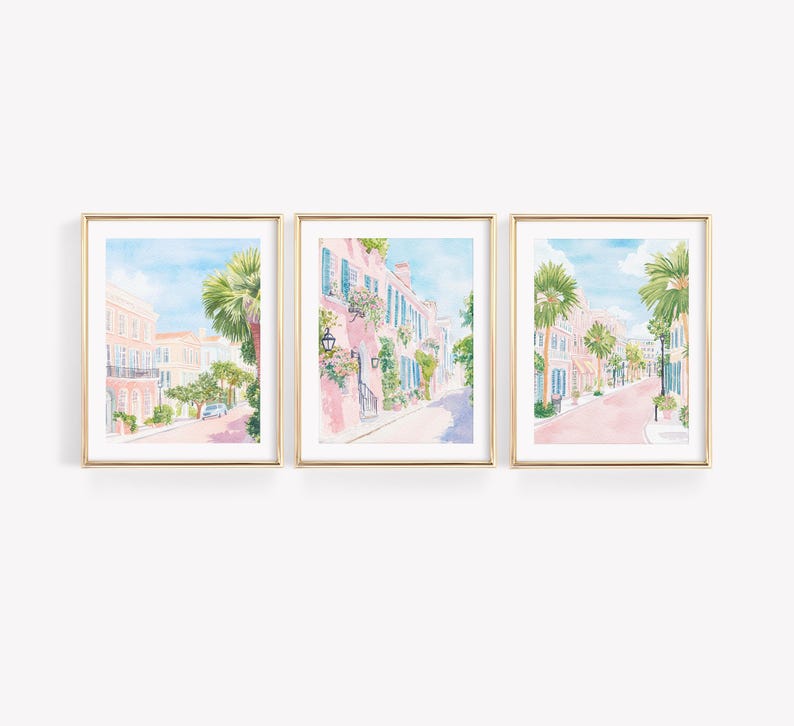 May include: Set of three framed watercolor prints depicting a charming street scene with pink buildings, palm trees, and a blue sky. The gold frames add an elegant touch to the artwork, perfect for home decor.