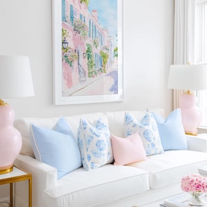 May include: A framed watercolor painting of a pink building with blue shutters and flowers hangs above a white sofa. The sofa is adorned with light blue and pink pillows. Two pink lamps with white shades flank the sofa.