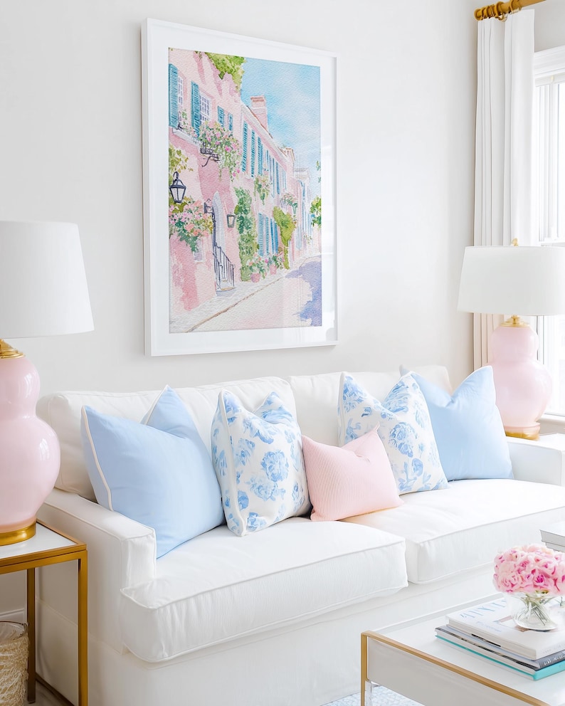 May include: A framed watercolor painting of a pink building with blue shutters and flowers hangs above a white sofa. The sofa is adorned with light blue and pink pillows. Two pink lamps with white shades flank the sofa.