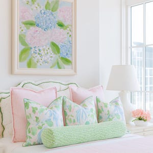 May include: A bed featuring a light pink striped coverlet, two large pink gingham pillows, three square pillows with a floral pattern in pink, blue, and green, and a long cylindrical bolster pillow in a green and white dotted pattern.