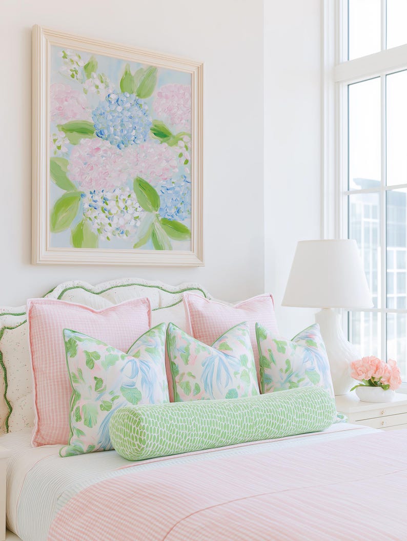 May include: A bed featuring a light pink striped coverlet, two large pink gingham pillows, three square pillows with a floral pattern in pink, blue, and green, and a long cylindrical bolster pillow in a green and white dotted pattern.