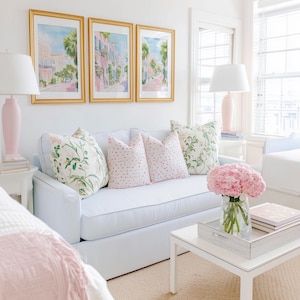 May include: A bright, airy living room scene featuring a light blue striped sofa adorned with floral and polka-dot pillows. Above the sofa, three framed watercolor paintings depict a street scene. A white coffee table holds a tray with books and a vase of pink flowers.