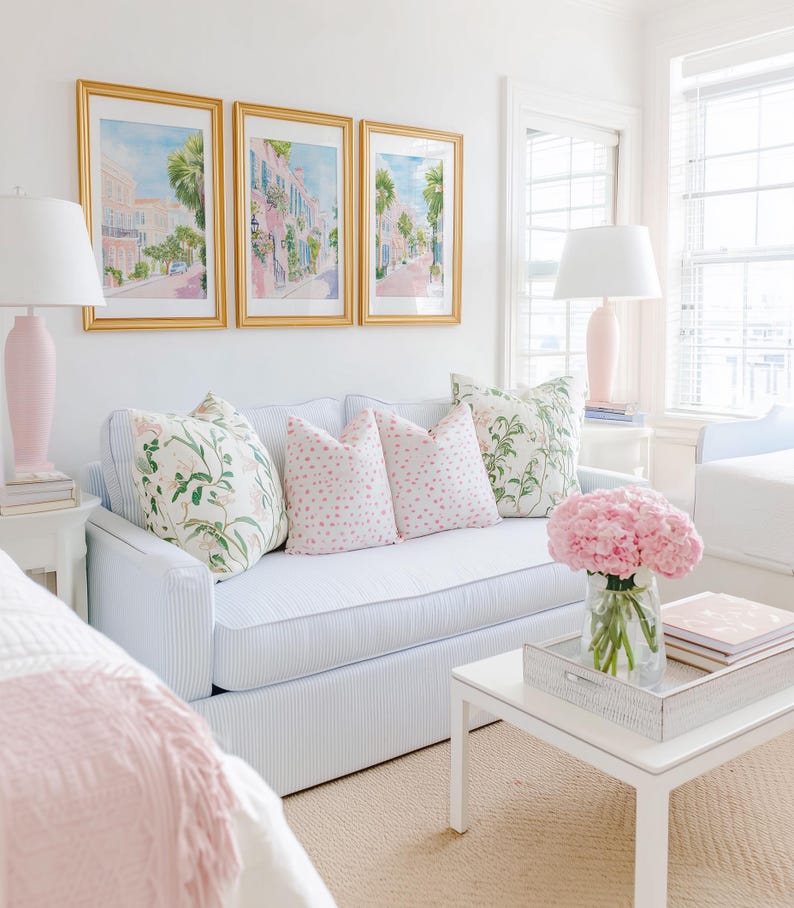 May include: A bright, airy living room scene featuring a light blue striped sofa adorned with floral and polka-dot pillows. Above the sofa, three framed watercolor paintings depict a street scene. A white coffee table holds a tray with books and a vase of pink flowers.