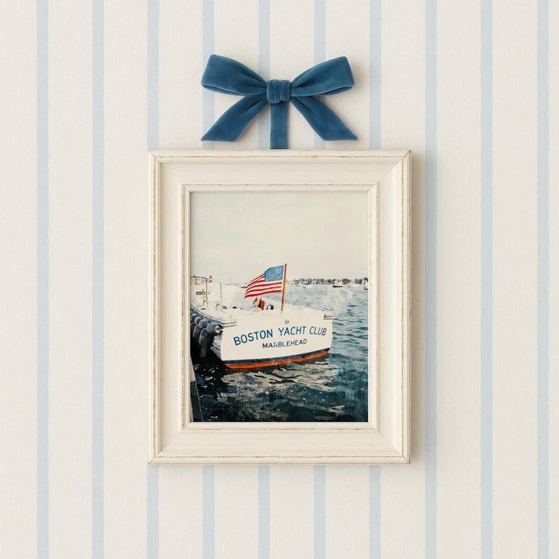 May include: A framed print of a boat with an American flag, labeled "Boston Yacht Club Marblehead." The white frame is topped with a blue velvet bow. The background is a light blue and white striped wall.