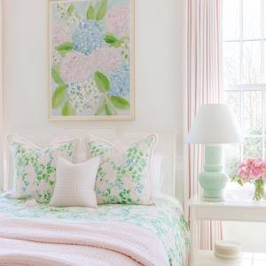 May include: A bedroom scene featuring a framed painting of blue and pink hydrangeas, two floral patterned throw pillows with pink piping, a small white gingham pillow, a green and white floral quilt, and a light pink textured blanket.