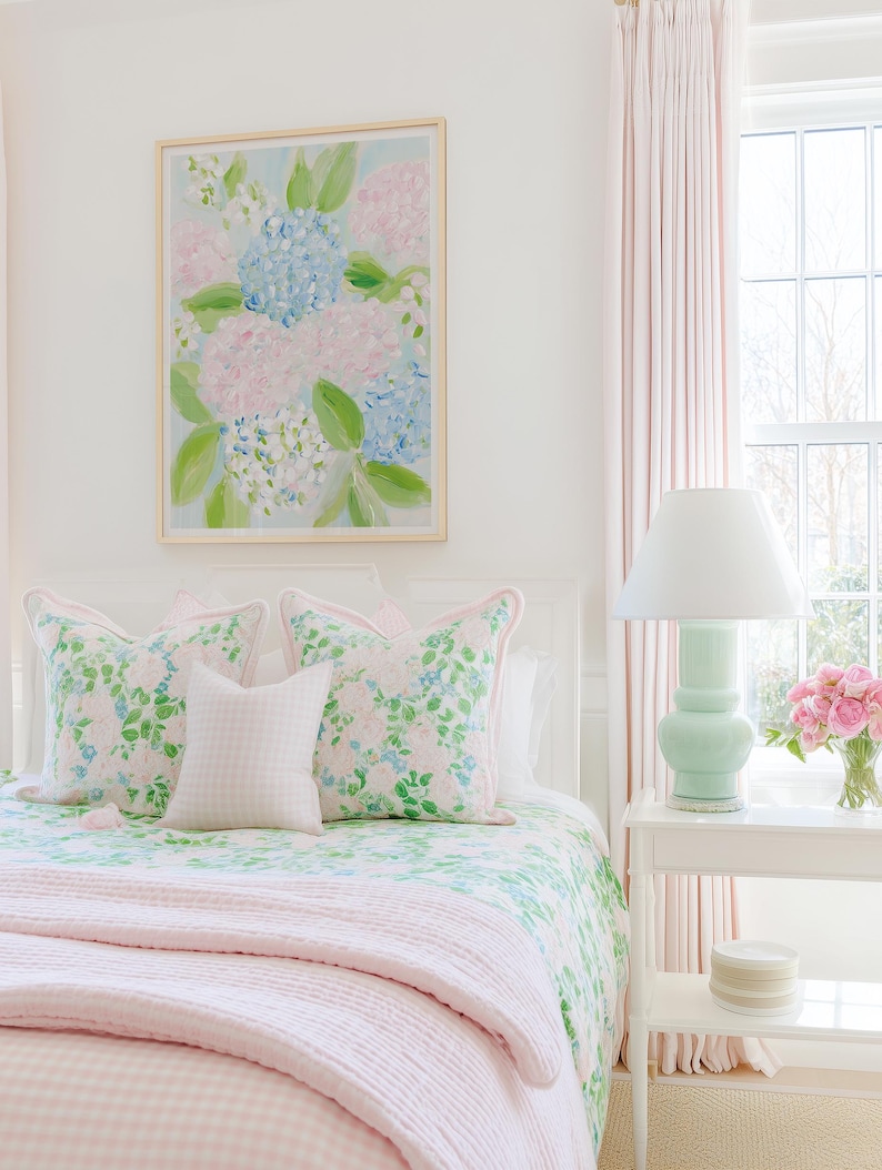 May include: A bedroom scene featuring a framed painting of blue and pink hydrangeas, two floral patterned throw pillows with pink piping, a small white gingham pillow, a green and white floral quilt, and a light pink textured blanket.