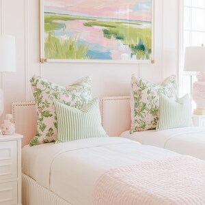 Op de afbeelding: Een slaapkamer met twee eenpersoonsbedden met lichtroze gestoffeerde hoofdeinden, wit beddengoed en lichtroze getextureerde plaids. De bedden zijn gestyled met kussens met bloemenprint en groen-wit gestreepte sierkussens onder een landschapsschilderij.