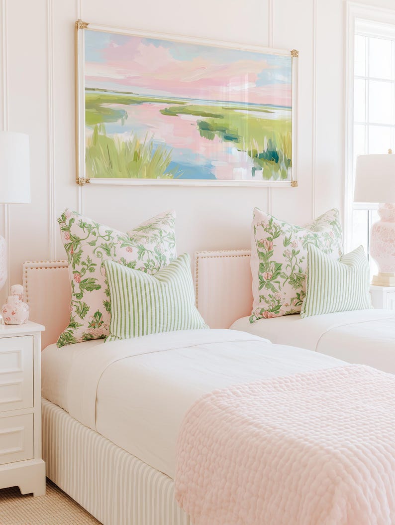 Op de afbeelding: Een slaapkamer met twee eenpersoonsbedden met lichtroze gestoffeerde hoofdeinden, wit beddengoed en lichtroze getextureerde plaids. De bedden zijn gestyled met kussens met bloemenprint en groen-wit gestreepte sierkussens onder een landschapsschilderij.