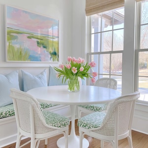 Op de afbeelding: Een witte ronde tafel met een centrale voet, twee witte stoelen met een rieten rugleuning en groen gedessineerde kussens, een ingebouwde bank met lichtblauwe kussens en een glazen vaas met roze tulpen onder een ingelijst landschapsschilderij.