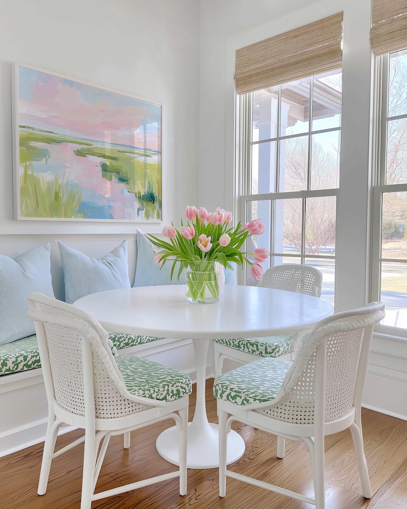 Op de afbeelding: Een witte ronde tafel met een centrale voet, twee witte stoelen met een rieten rugleuning en groen gedessineerde kussens, een ingebouwde bank met lichtblauwe kussens en een glazen vaas met roze tulpen onder een ingelijst landschapsschilderij.