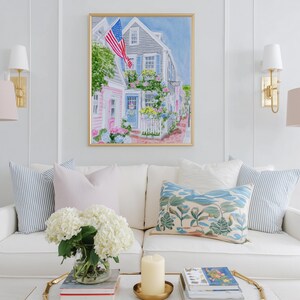 May include: A framed watercolour painting of a coastal street scene with an American flag. The artwork hangs above a white sofa with decorative cushions. The room is decorated with gold lamps and a coffee table with a candle and flowers.