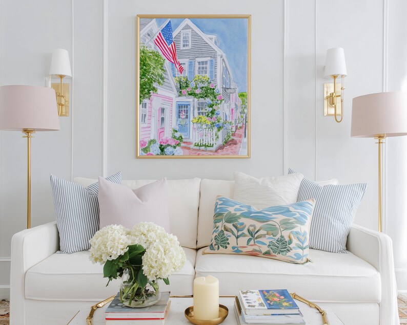 May include: A framed watercolour painting of a coastal street scene with an American flag. The artwork hangs above a white sofa with decorative cushions. The room is decorated with gold lamps and a coffee table with a candle and flowers.