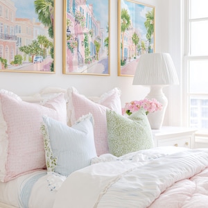 May include: A bedroom scene featuring a bed with pink and blue pillows and a white comforter. Above the bed are three framed watercolor paintings of a street scene. A white lamp and vase of pink flowers sit on a white nightstand.