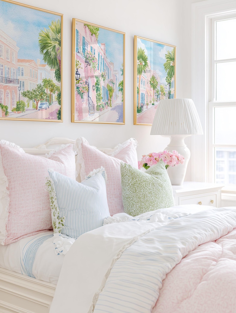 May include: A bedroom scene featuring a bed with pink and blue pillows and a white comforter. Above the bed are three framed watercolor paintings of a street scene. A white lamp and vase of pink flowers sit on a white nightstand.