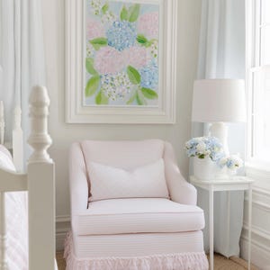 May include: A pale pink upholstered armchair with a matching lumbar pillow and a decorative fringe skirt at the base. The chair is positioned next to a white side table holding a white lamp and a vase filled with blue and white hydrangea flowers.