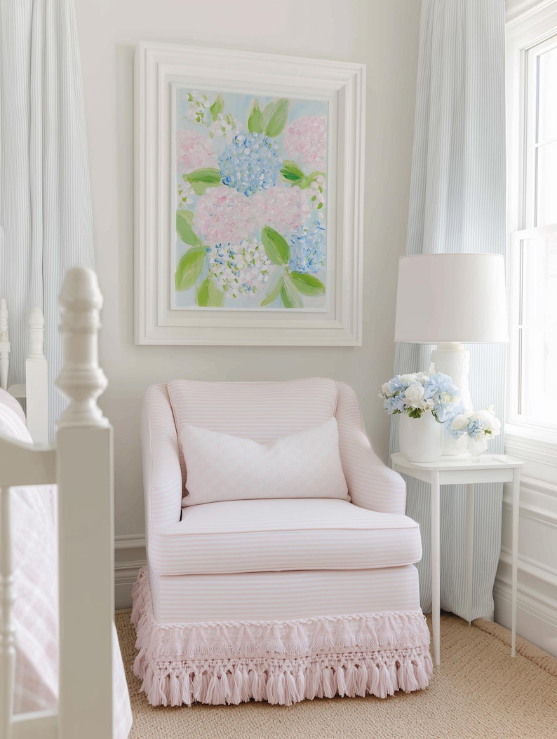 May include: A pale pink upholstered armchair with a matching lumbar pillow and a decorative fringe skirt at the base. The chair is positioned next to a white side table holding a white lamp and a vase filled with blue and white hydrangea flowers.