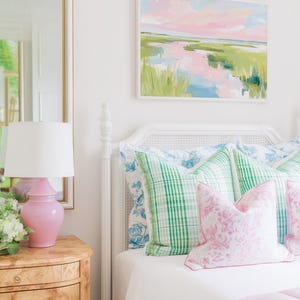 Op de afbeelding: Een wit bedframe met een rieten hoofdbord, opgemaakt met groen geruite kussens en roze kussens met bloemenprint. Een roze keramische lamp staat op een nachtkastje van wortelhout naast een spiegel. Een landschapsschilderij hangt boven het bed.