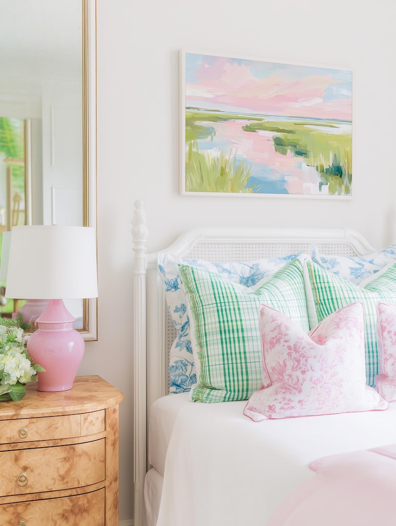 Op de afbeelding: Een wit bedframe met een rieten hoofdbord, opgemaakt met groen geruite kussens en roze kussens met bloemenprint. Een roze keramische lamp staat op een nachtkastje van wortelhout naast een spiegel. Een landschapsschilderij hangt boven het bed.