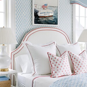 May include: A bedroom scene featuring a white headboard with red trim, adorned with white pillows and patterned throw pillows. A framed picture of a boat with an American flag hangs above the bed. The room has blue patterned wallpaper and matching window treatments.