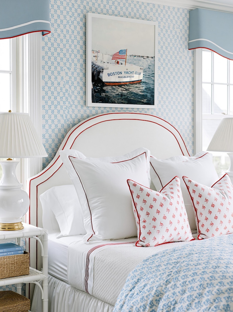 May include: A bedroom scene featuring a white headboard with red trim, adorned with white pillows and patterned throw pillows. A framed picture of a boat with an American flag hangs above the bed. The room has blue patterned wallpaper and matching window treatments.