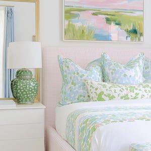 Op de afbeelding: Een slaapkamer met een roze geruit gestoffeerd hoofdeinde, wit beddengoed met een groen en blauw bloemmotief en bijpassende kussens. Op een wit nachtkastje staat een groene keramische lamp met een witte kap naast een spiegel met een gouden lijst.