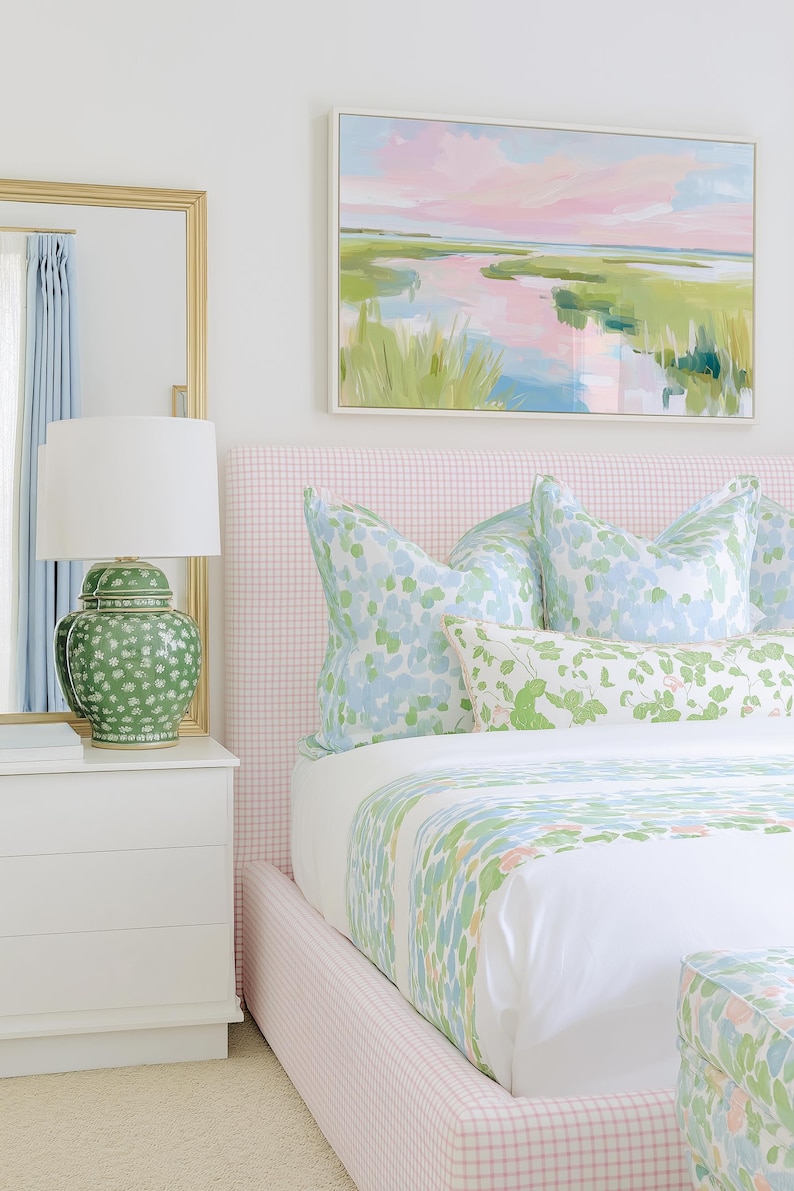 Op de afbeelding: Een slaapkamer met een roze geruit gestoffeerd hoofdeinde, wit beddengoed met een groen en blauw bloemmotief en bijpassende kussens. Op een wit nachtkastje staat een groene keramische lamp met een witte kap naast een spiegel met een gouden lijst.