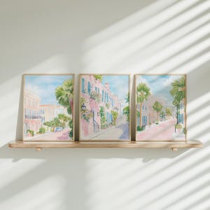 May include: Three framed watercolor prints depicting a street scene with pink buildings, palm trees, and blue shutters. The artwork is displayed on a light-colored wooden shelf against a white wall. The style is reminiscent of a coastal town.