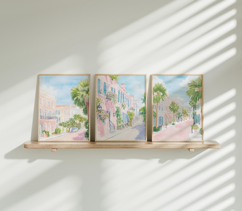 May include: Three framed watercolor prints depicting a street scene with pink buildings, palm trees, and blue shutters. The artwork is displayed on a light-colored wooden shelf against a white wall. The style is reminiscent of a coastal town.