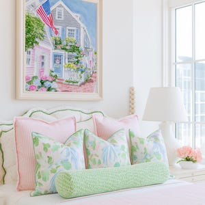 May include: A bedroom scene with a bed dressed in pink and white striped bedding, complemented by pink and patterned pillows. A green bolster cushion provides contrast. Above the bed is a framed watercolour painting of a coastal town with an American flag.