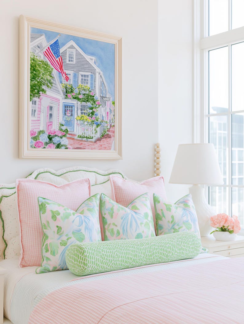 May include: A bedroom scene with a bed dressed in pink and white striped bedding, complemented by pink and patterned pillows. A green bolster cushion provides contrast. Above the bed is a framed watercolour painting of a coastal town with an American flag.