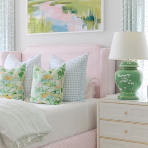 Op de afbeelding: Een slaapkamer met een roze en wit geruit gestoffeerd bedframe, twee lichtblauw geruite kussens, twee kussens met bloemenprint, een witte lamp met een groene keramische voet en een wit nachtkastje met drie lades en gouden accenten.