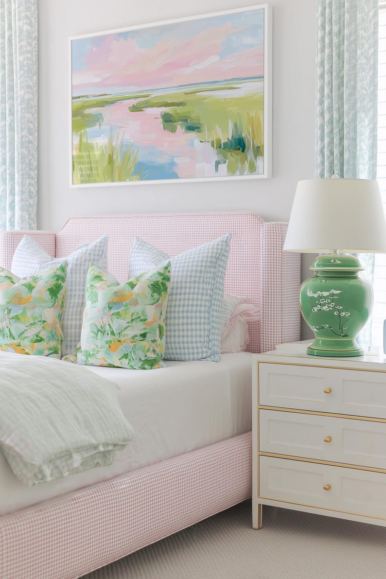 Op de afbeelding: Een slaapkamer met een roze en wit geruit gestoffeerd bedframe, twee lichtblauw geruite kussens, twee kussens met bloemenprint, een witte lamp met een groene keramische voet en een wit nachtkastje met drie lades en gouden accenten.