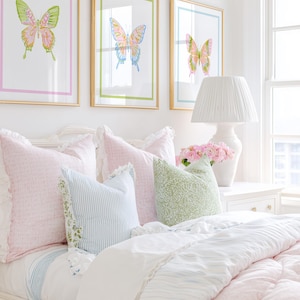 May include: A bedroom scene featuring three framed butterfly art prints above a bed with pink and blue pillows and a white comforter. A white lamp and vase of pink flowers sit on the nightstand.