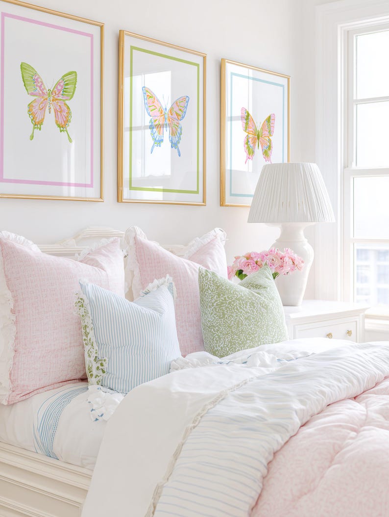 May include: A bedroom scene featuring three framed butterfly art prints above a bed with pink and blue pillows and a white comforter. A white lamp and vase of pink flowers sit on the nightstand.