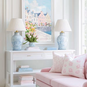 Puede incluir: Una escena de sal&oacute;n luminosa y espaciosa con una consola blanca con dos l&aacute;mparas azules y blancas con pantallas plisadas. Un sof&aacute; rosa con cojines estampados est&aacute; cerca. Una pintura enmarcada de un pueblo costero cuelga sobre la mesa, y un jarr&oacute;n de tulipanes est&aacute; sobre la mesa.