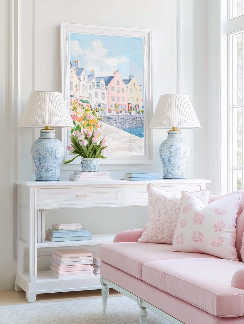 Puede incluir: Una escena de sal&oacute;n luminosa y espaciosa con una consola blanca con dos l&aacute;mparas azules y blancas con pantallas plisadas. Un sof&aacute; rosa con cojines estampados est&aacute; cerca. Una pintura enmarcada de un pueblo costero cuelga sobre la mesa, y un jarr&oacute;n de tulipanes est&aacute; sobre la mesa.