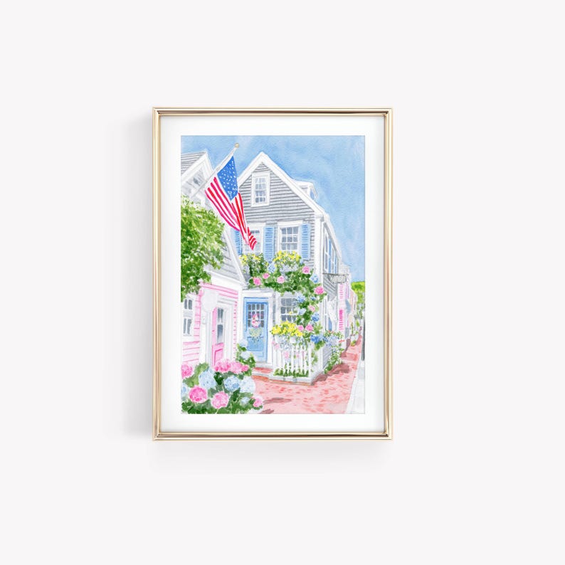 May include: Watercolour print depicting a quaint street scene with vibrant houses, decorated with flowers and an American flag. The houses are painted in pink, grey, and blue, with a brick pathway. The artwork is framed in a gold frame.