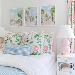 May include: A bedroom scene featuring a bed with a pink headboard, adorned with floral and gingham pillows. Above the bed, three framed watercolor prints depict a street scene. A pink lamp and floral curtains complete the decor.