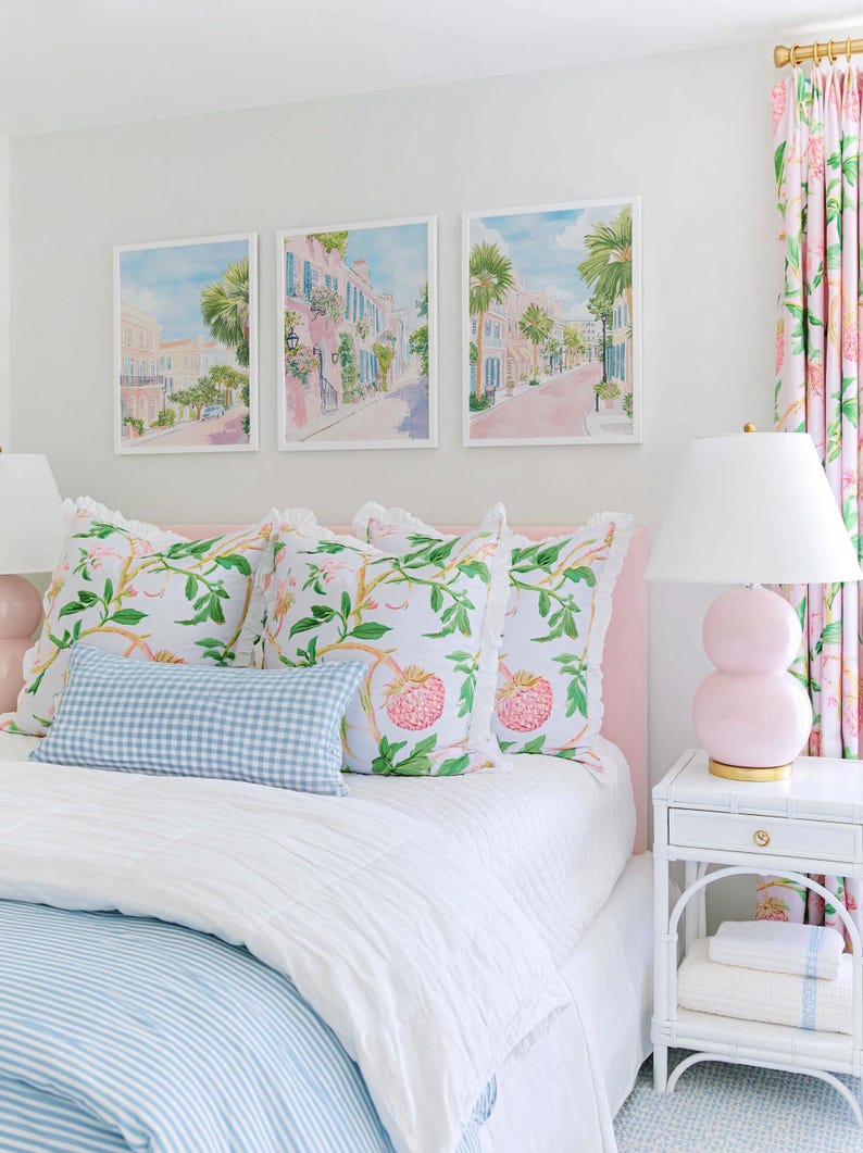 May include: A bedroom scene featuring a bed with a pink headboard, adorned with floral and gingham pillows. Above the bed, three framed watercolor prints depict a street scene. A pink lamp and floral curtains complete the decor.