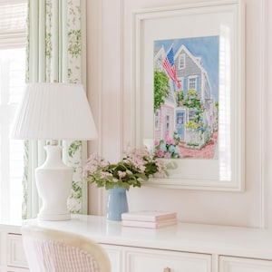 May include: A white desk with a white lamp, a blue vase with flowers, and a framed watercolour painting of a house with an American flag. The painting is the focal point, with a pink and blue colour scheme.