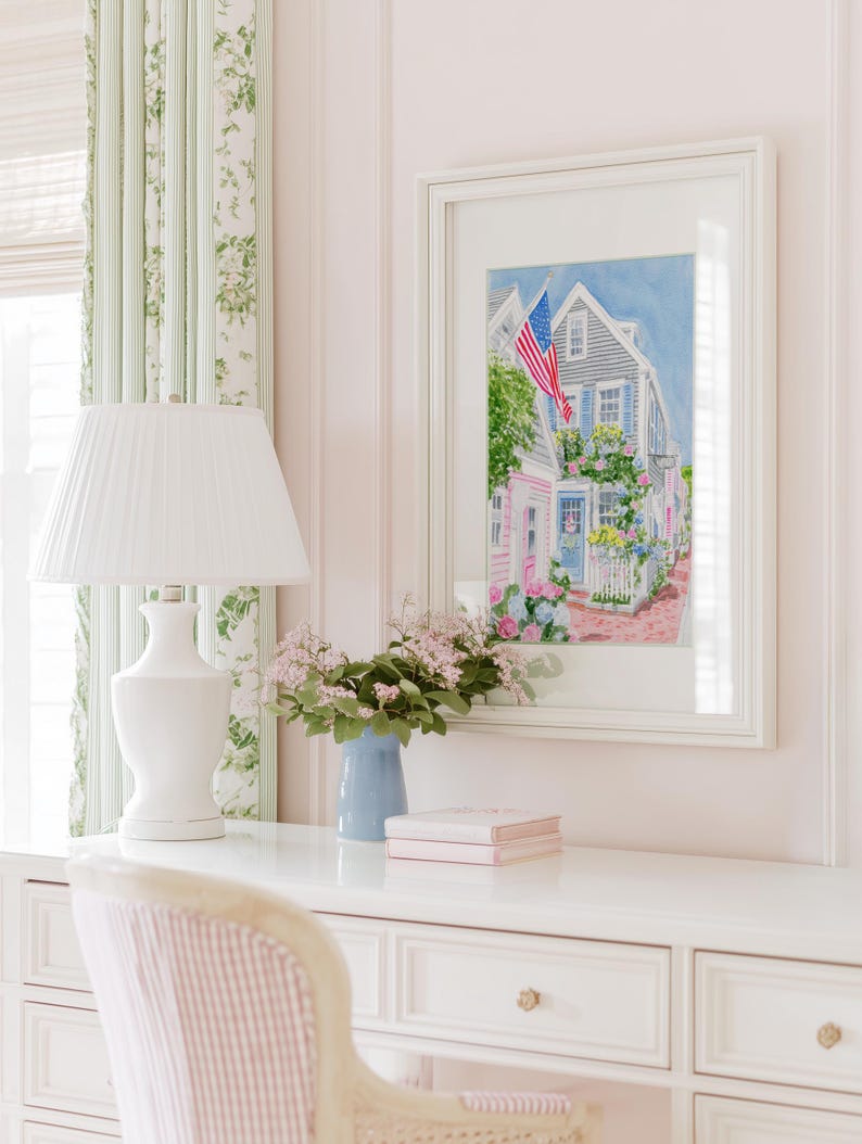 May include: A white desk with a white lamp, a blue vase with flowers, and a framed watercolour painting of a house with an American flag. The painting is the focal point, with a pink and blue colour scheme.