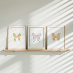 May include: Three framed butterfly art prints. Each print features a watercolor butterfly with pastel colors, set against a white background. The frames have colored borders in pink, green, and blue. The prints are displayed on a wooden shelf.