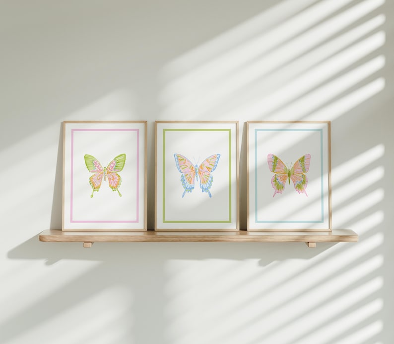 May include: Three framed butterfly art prints. Each print features a watercolor butterfly with pastel colors, set against a white background. The frames have colored borders in pink, green, and blue. The prints are displayed on a wooden shelf.