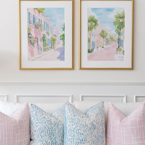 May include: Two framed watercolor prints of a street scene with pink buildings and blue shutters, hanging above a white couch with decorative pillows. The pillows are in shades of pink and blue.